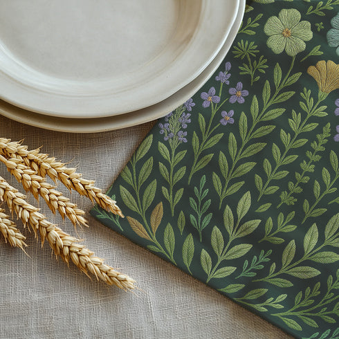 A 4-piece cloth napkin set featuring embroidered-style botanical illustrations of wildflowers and herbs in shades of gold, mint, and lavender on a dark green background; made from soft polyester, square-shaped, with a luxe and mystical design.